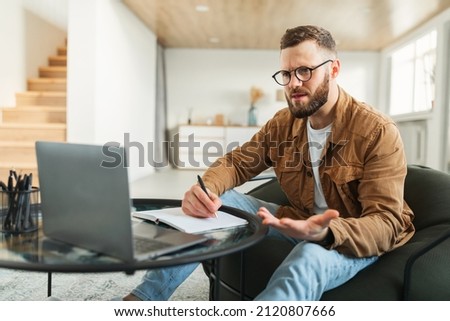 Discontented Businessman Using Laptop Taking Notes Having Problems With Accounting Working Online Sitting In Chair At Home, Wearing Eyeglasses. Distance Work Issues. Selective Focus