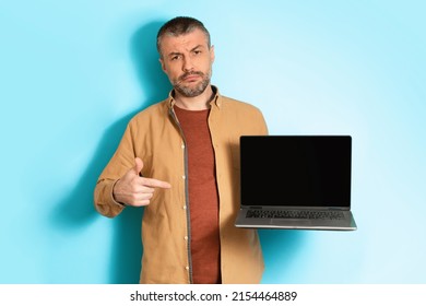 Discontented Middle Aged Man Showing Laptop Pointing Finger At Empty Screen Frowning Looking At Camera Disapproving Bad Website Standing Over Blue Background. Studio Shot, Mockup