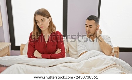 A discontented woman and a man sit apart in bed, symbolizing relationship issues in a bedroom setting.