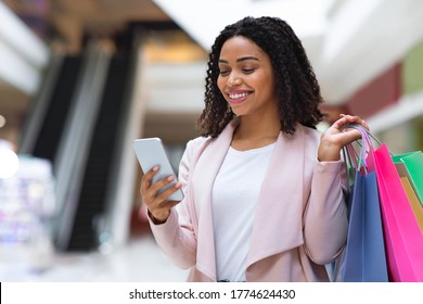 Discount App. Smiling African American Woman With Shopping Bags Using Smartphone In Mall, Checking Sale Offers, Free Space