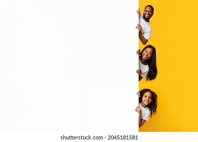 Discount. Smiling young black family of three holding white advertisement board, presenting free copy space for your text or design, positive people peeking out blank sign board banner, yellow studio - Powered by Shutterstock