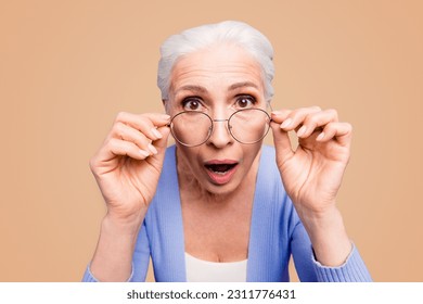 Discounts and sales What a great news Close up portrait of old woman looks over her spectacles in surprise, opening her mouth and eyes wide isolated on shine purple background