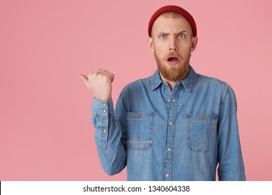 Discouraged amazed man, opened his mouth in surprise and points with thumb to the left side on copy space, guy in red hat with red thick beard, wears denim shirt,isolated on pink background