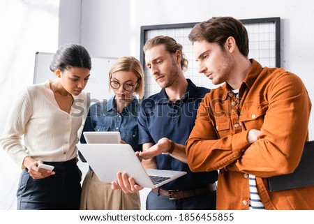 discouraged man pointing with finger at laptop near dissatisfied multicultural colleagues