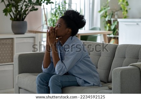 Discouraged pensive African American woman looking into distance thinking about ways to solve financial problems. Frightened black girl sits on sofa in living room having lost mood due to inflation
