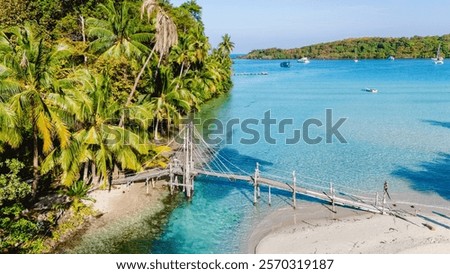 Discover the beauty of Koh Kood in Thailand, where a charming wooden bridge connects sandy shores. Lush palm trees sway gently as the sun sets over tranquil waters, painting a picturesque view.