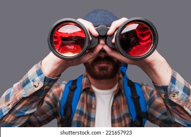 Discovering new places. Confident young bearded man carrying backpack and looking through binoculars while standing against grey background