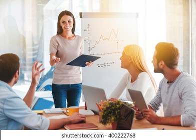 Discussing new business ideas. Cheerful young woman standing near whiteboard and smiling while her colleagues sitting at the desk 