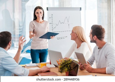 Discussing some business issues. Cheerful young woman standing near whiteboard and smiling while her colleagues sitting at the desk 