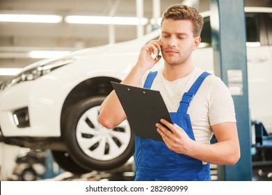 Discussing Some Car Problems. Handsome Young Man Talking On The Mobile Phone And Looking At Clipboard While Standing In Workshop With Car In The Background