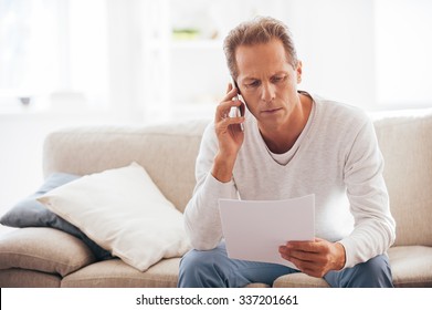 Discussing That Document. Serious Mature Man Holding Paper And Talking On The Mobile Phone While Sitting On The Couch At Home