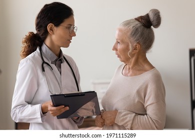 Discussing Therapy. Serious Aged Woman Clinic Visitor Stand Close To Confident Female Doctor With Clipboard Ask Questions About Diagnosis. Old Lady Patient Talking With Young Gp Specify Prescription