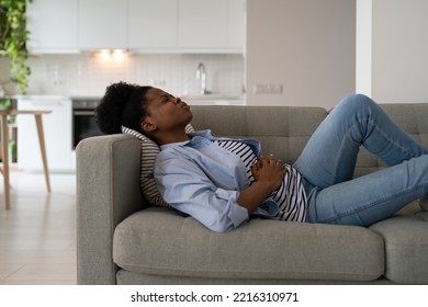 Diseased African American Woman Holding Stomach Feeling Pain And Anguish After Alimentary Poisoning Or Junk Food. Suffering Girl Lies On Couch Needs Trip To Doctor Gastroenterologist Or Nutritionist