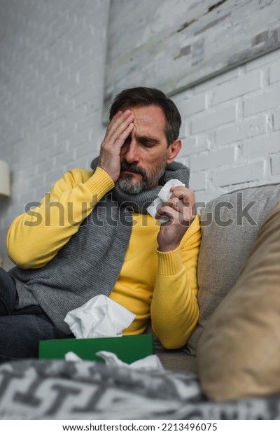 diseased man obscuring\
face with hand while sitting on couch with paper napkin and\
suffering from\
headache