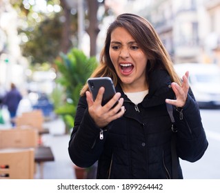 Disgruntled Emotional Young Hispanic Woman Looking At Phone Screen And Screaming During Walk Along Autumn City