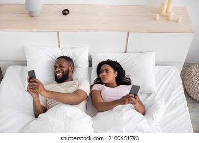 Disgruntled millennial african american wife looks at husband smartphone on bed in bedroom interior, top view. Gadget addiction, online games and social networks, treason and relationship problems