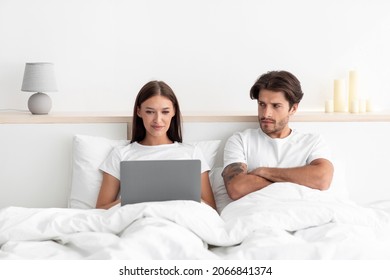 Disgruntled Sad Offended Caucasian Husband With Crossed Arms Looks At Wife, Typing On Laptop In Bed In White Bedroom Interior. Covid-19 Pandemic, Jealousy, Work At Home, Social Networks, Free Space