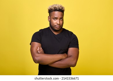 A Disgruntled Serious Black Man Stands In A Closed Position, With His Hands Folded On His Chest Cross To Cross. A Strong Guy With Big Muscles Stands In A T-shirt On A Yellow Background And Is Sad