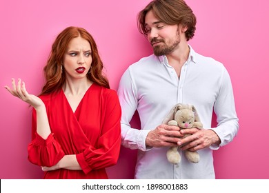 disgruntled woman shocked by immature man holding toy in hands, he is in love with her, man and woman in party clothes. isolated on pink background - Shutterstock ID 1898001883