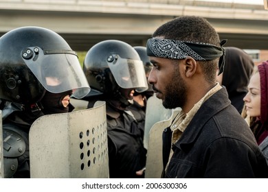 Disgruntled Young Black Bearded Man In Bandana Standing In Front Of Police Officers With Riot Helmets And Shields Outdoors