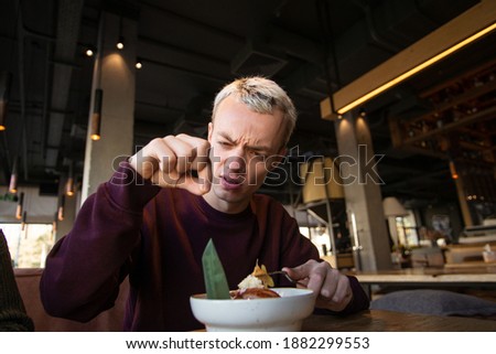 Disgusted young man found hair in his meal at cafe and looks at it with loathing. Bad customer service concept. Blond young man in casual clothes isn't satisfied with the food.