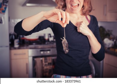 A disgusted young woman is holding a dead mouse by it's tail in the kitchen