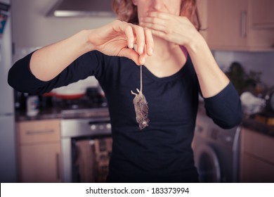 A disgusted young woman is holding a dead mouse by it's tail in the kitchen