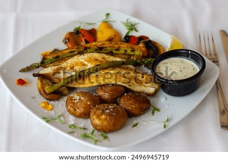 A dish featuring baked fish, roasted potatoes, grilled vegetables, and a sauce. Un plato con pescado al horno, papas asadas, verduras a la parrilla y una salsa.