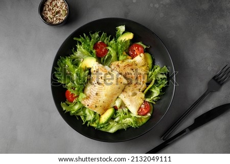 Dish with Gourmet fried halibut fish on Vegetable, fresh green salad. Top view. Black plate. Fork, knife