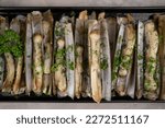 Dish of razor clam grilled with garlic, parsley, olive oil and lemon slice on gray stone table.