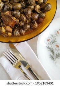 Dish Of Snails With Pork Meat And Sauce Served On The Table From Northern Spain, Catalonia. White Place Setting For One Person With Plate, Knife And Fork. 
