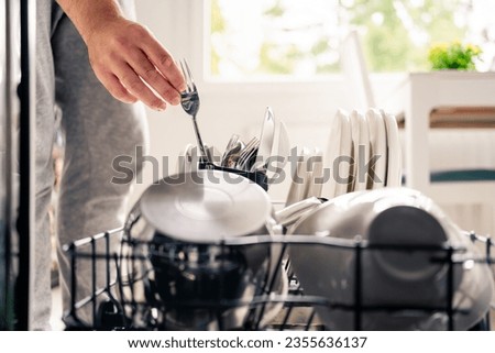 Dish washer machine in kitchen. Man loading dishwasher. Washing plates. Fork in hand. Full of cutlery. Clean or dirty. Pot and tableware. Household chores and housework in family. Unloading utensils.