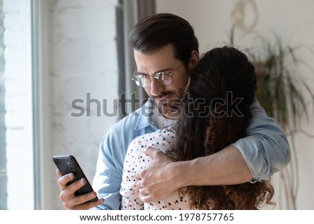 Dishonest young man cuddling loving woman, using smartphone behind back, checking her calls or communicating with mistress. Unfair millennial husband cheating wife, betrayal lack of trust concept.