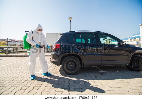 Disinfector in a protective suit\
and mask sprays disinfectants of car outdoors. Coronavirus\
Pandemic.