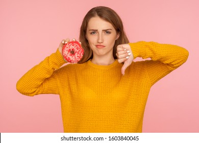 Dislike To Junk Food. Portrait Of Displeased Red Hair Girl In Sweater Showing Thumbs Down Gesture And Holding Sweet Donut, Harmful Sugary Confectionery, Diabetes. Indoor Studio Shot, Pink Background