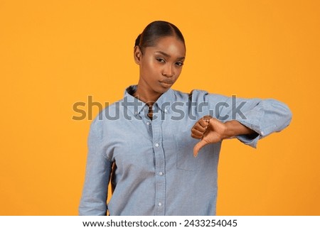 Dislike. Young black woman expresses discontent giving thumbs down gesture in studio with orange background, conveying negative or disapproving concept in modern advertising