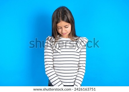 Dismal gloomy rejected beautiful kid girl wearing striped t-shirt over blue background has problems and difficulties, curves lower lip and closes eyes in despair, being in depression