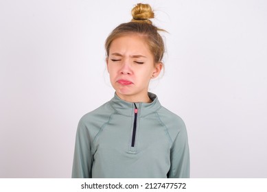 Dismal Gloomy Rejected Little Caucasian Kid Girl With Hair Bun Wearing Technical Shirt Over White Background  Has Problems And Difficulties, Curves Lower Lip And Closes Eyes In Despair,