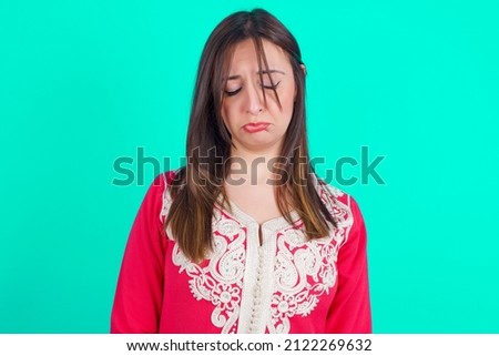 Dismal gloomy rejected young beautiful moroccan woman wearing traditional caftan dress over green background has problems and difficulties, curves lower lip and closes eyes in despair.