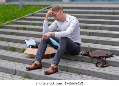 Dismissal Of An Employee Due To A Coronavirus Epidemic. A Man Sits On The Steps. Next To Him Is His Stationery. He Has A Medical Mask In His Hand.