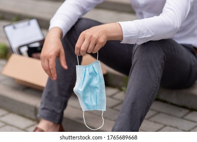 Dismissal Of An Employee Due To A Coronavirus Epidemic. A Man Sits On The Steps. Next To Him Is His Stationery. He Has A Medical Mask In His Hand.