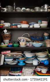 Disorganised kitchen cupboard with stacks of mismatched ceramic plates and bowls, piles of mugs and glasses and lots of jars and vases randomly shoved onto the shelves