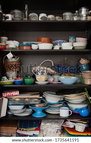 Disorganised kitchen cupboard with stacks of mismatched ceramic plates and bowls, piles of mugs and glasses and lots of jars and vases randomly shoved onto the shelves