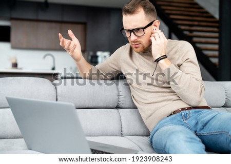 Disoriented young man looking at computer screen, holding the hand on the earphone, wrinkling the forehead, checking the connection, doesn't hear the companion, computer is broken, what happened
