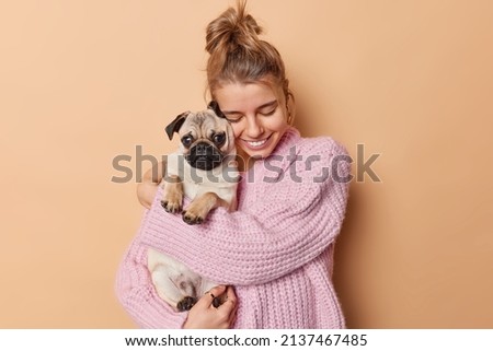 Display of affection. Happy young woman with combed hair embraces pug dog has fun being mom of puppy wears knitted sweater isolated over beige background. Female pet lover with domestic animal