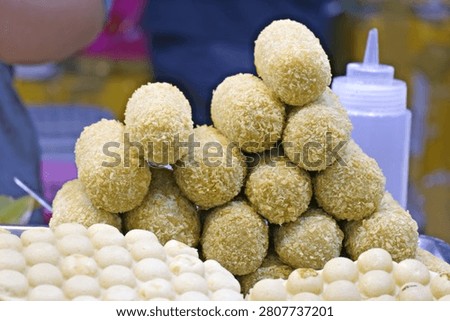 A display of golden deep-fried breaded snacks and miniature round cakes at a bustling street food market in Vinh Long, Vietnam.
