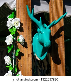 Display Of Teal Green Ceramic Deer Head Hanging Between Two Wooden Beams With A White Row Of Fake White Roses And Green Leaves Hanging Down On The Side Of Wooden Beam