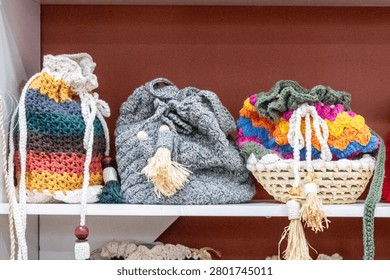 Display of three handmade crochet drawstring bags with straw and wooden details on a white shelf. Sharp technical focus and terracotta background.