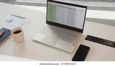 Displaying spreadsheet, open silver laptop at center of white desk, with coffee mug and smartphone. Computer, workspace, modern, minimalist, organized, analytics, productivity