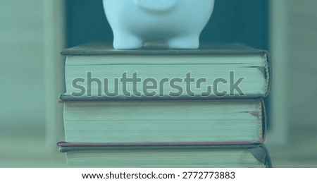 Displaying stack of three hardcover books with white ceramic piggy bank sitting on wooden table. Stilllife, literature, porcelain, desk, depthoffield, teal, beige